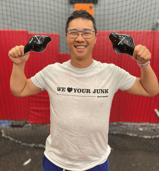 Smiling man in NuttyBuddy We ❤ Your Junk athletic fit shirt, holding black gear in a sports facility.