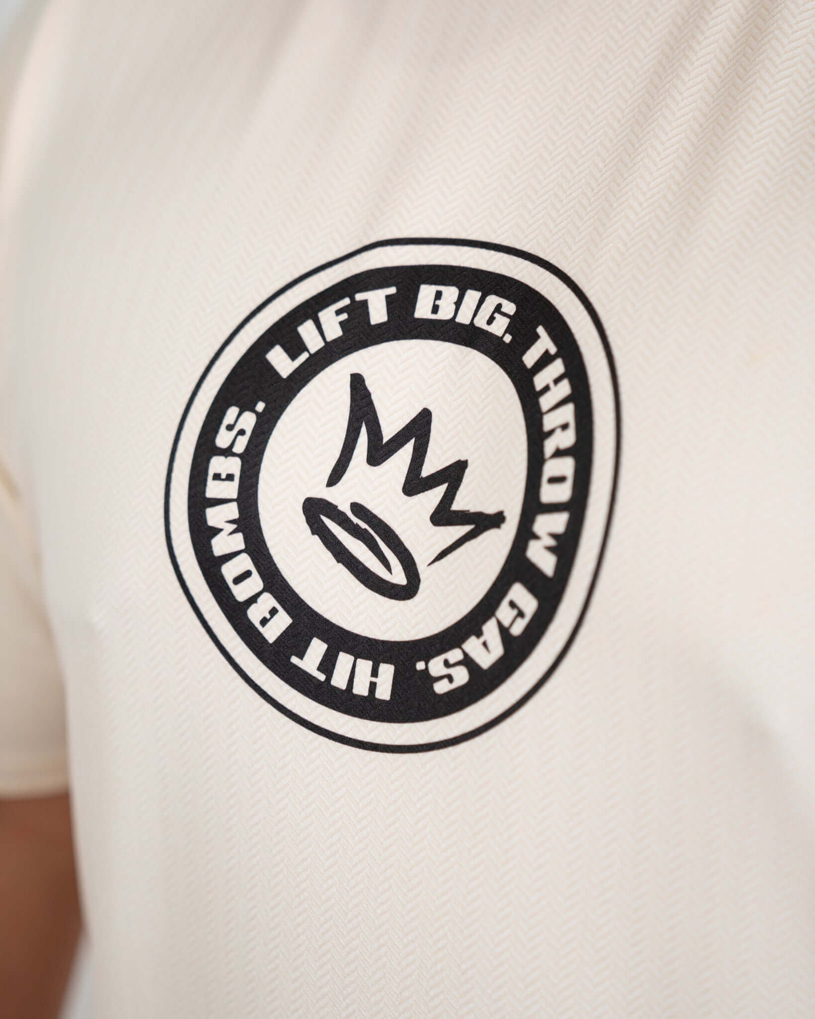 Close-up of King of Juco Tan Recovery Tee logo featuring the phrase 'Lift Big, Throw Bombs' in black on a beige background.