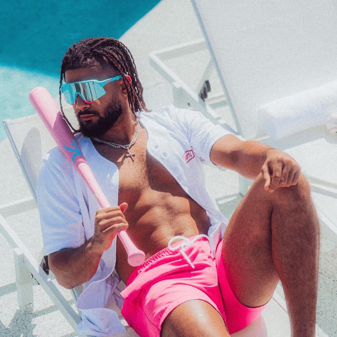 Man lounging by the pool wearing sunglasses, a white shirt, and pink shorts, holding a pink baseball bat.