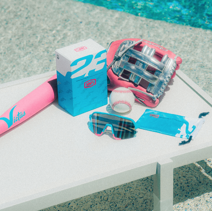 Blue and pink baseball accessories on a table near a pool, featuring a Victus bat, glove, baseball, and Speedcraft sunglasses.