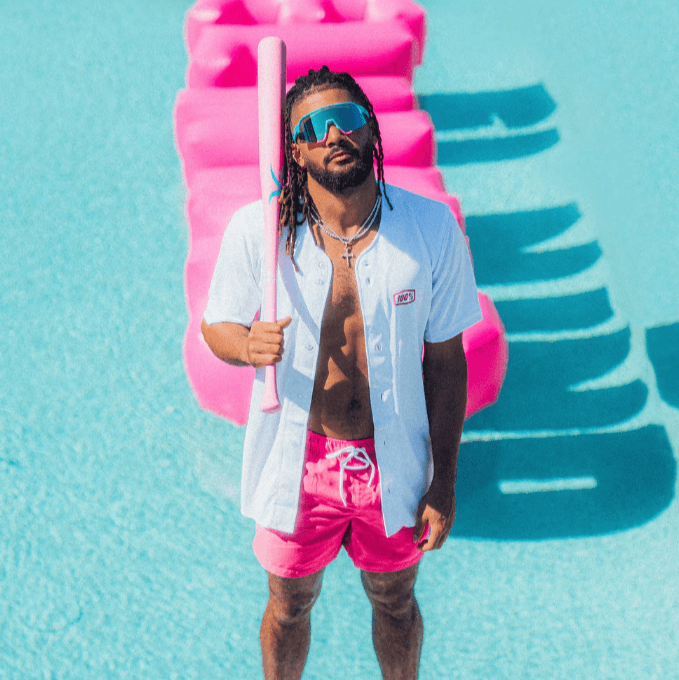 Man in sunglasses and pink shorts posing by a pool, holding a baseball bat with a pink slide in the background.