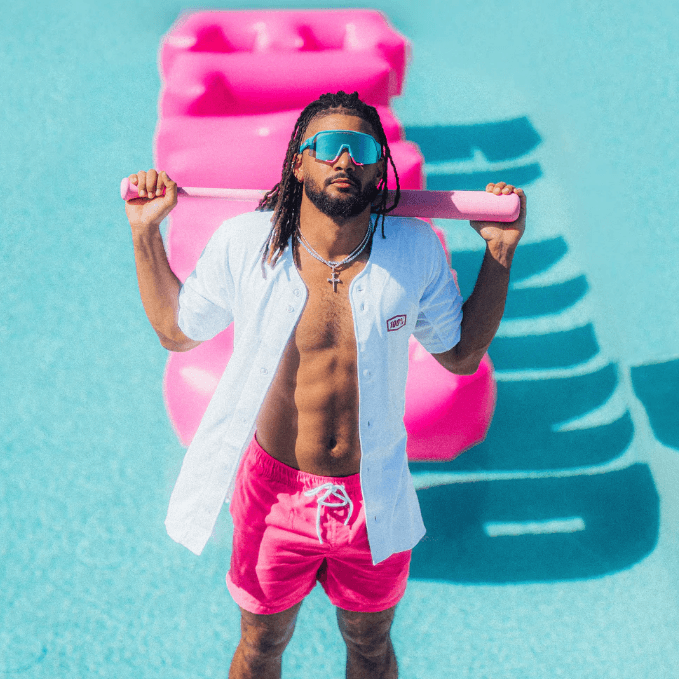 Man in sunglasses and pink swim trunks poses poolside with colorful floaties in the background, emphasizing summer vibes.