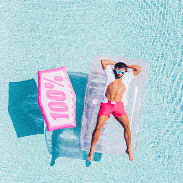 Man lounging on a pink inflatable float in a swimming pool, wearing sunglasses and shorts, enjoying summer vibes.
