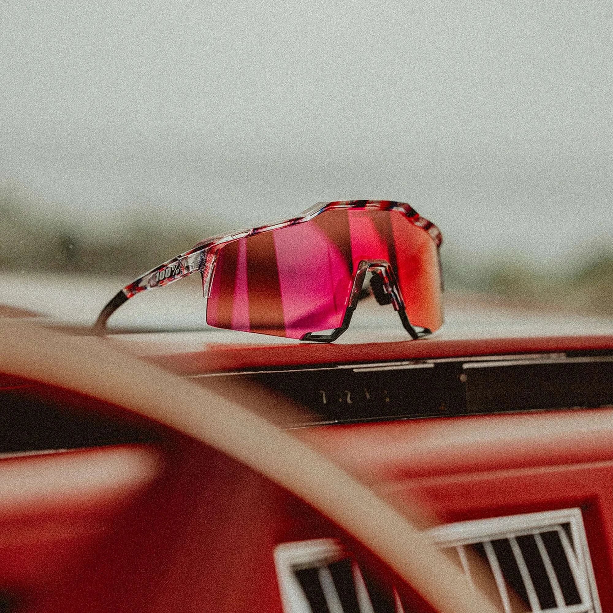 100% Speedcraft FDJ Gloss Translucent Red/Blue sunglasses with HiPER Fire Red Mirror Lens, side profile view on car dashboard.