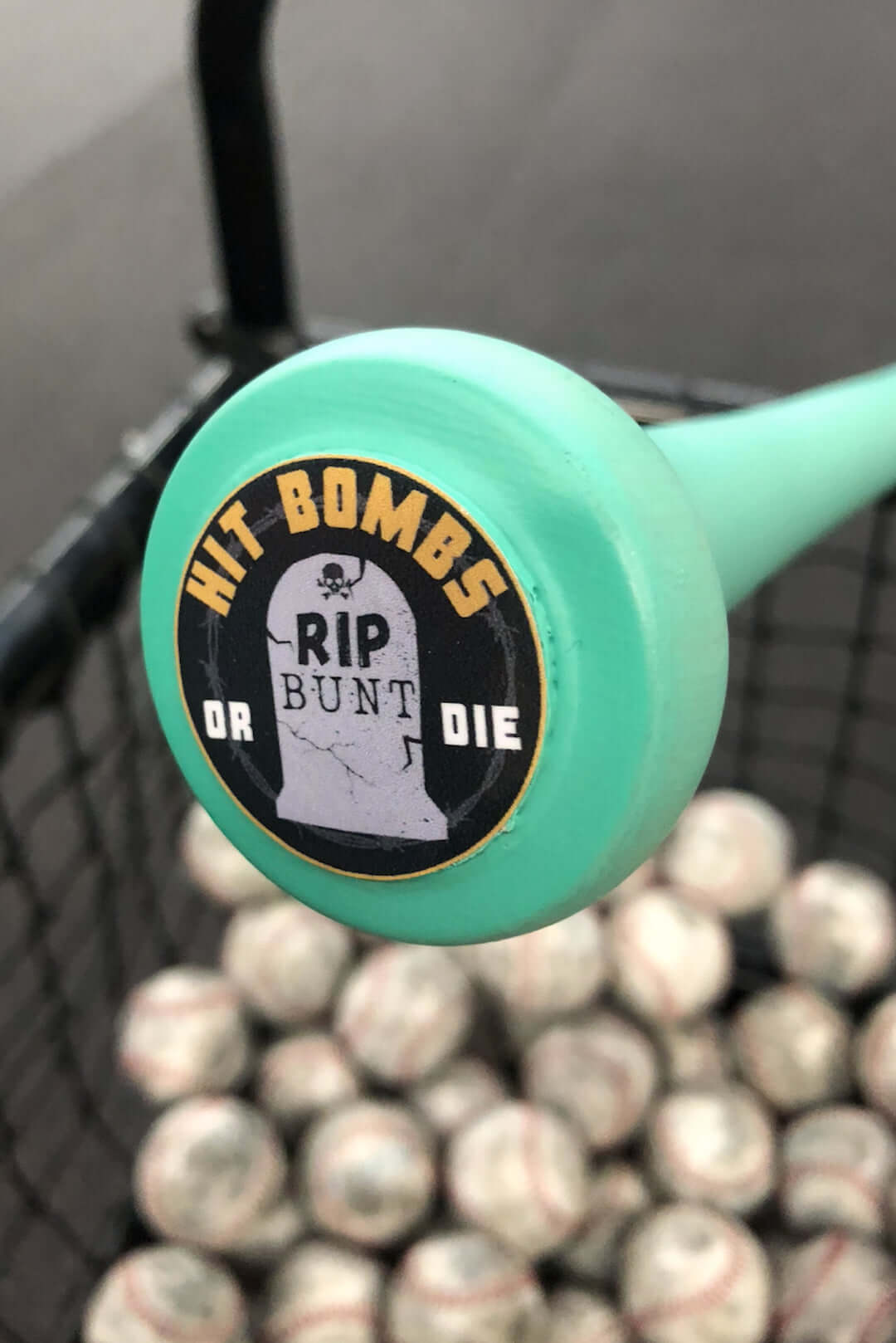 Knob of a matte mint wood bat with "Hit Bombs or Die" sticker, surrounded by baseballs in a practice basket.