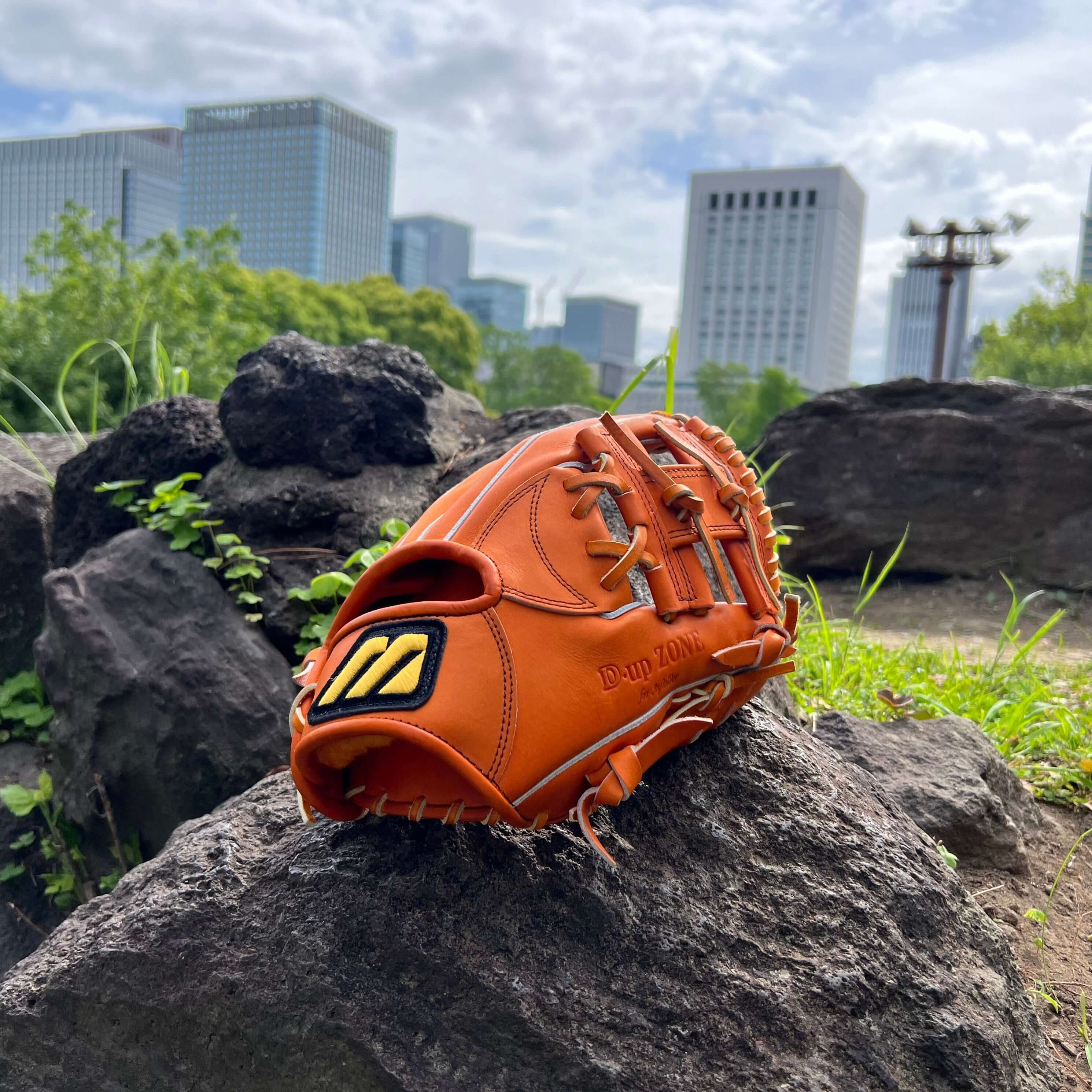 Mizuno Pro D-Up Zone July Limited Edition glove in orange displayed on rocks with city skyline in the background.