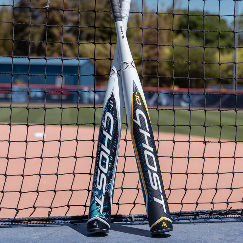 Two Easton Ghost Tie-Dye fastpitch bats leaning against a baseball field fence, showcasing their unique designs.