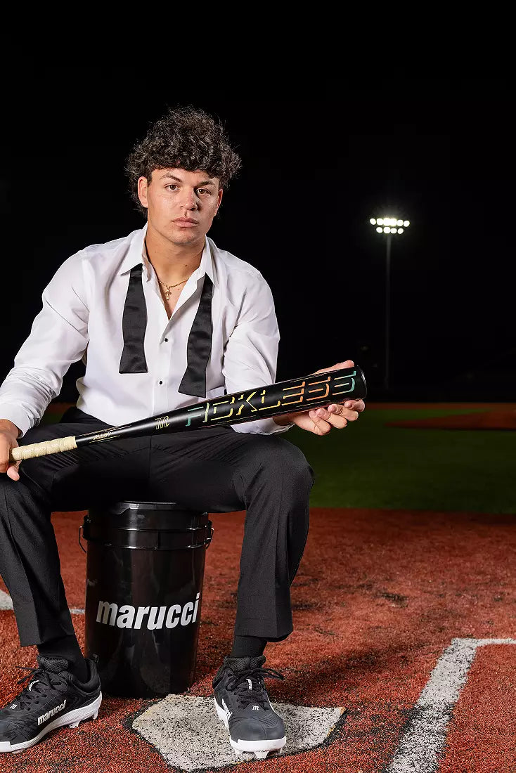 Baseball player sitting on a Marucci bucket holding a Marucci RCKLESS GEN 2 (-3) BBCOR Baseball Bat on a field at night.
