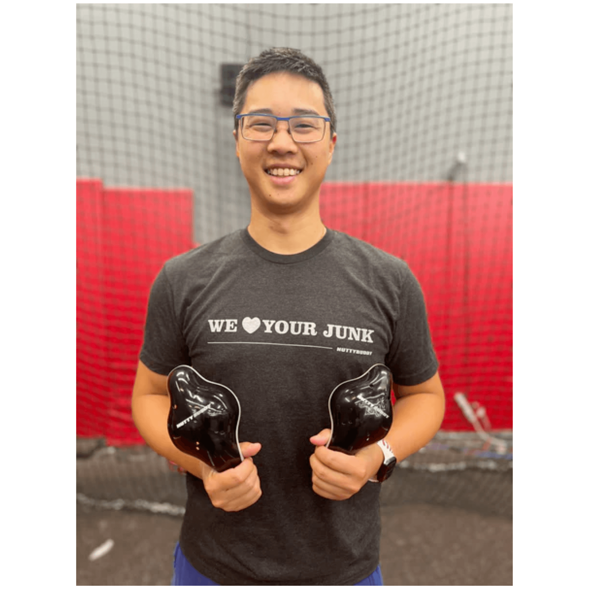Young man in We ❤ Your Junk athletic fit shirt, holding NuttyBuddy cups, smiles against a sports facility backdrop.
