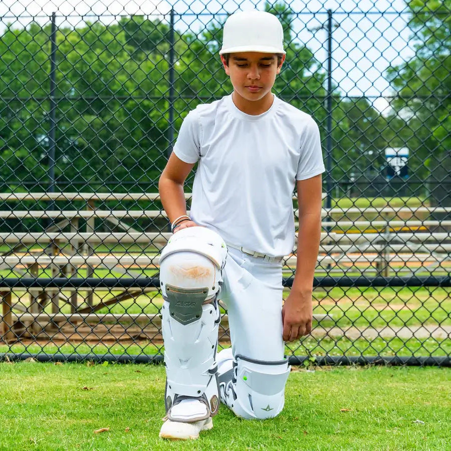 Young catcher kneeling on grass wearing All-Star MVP-PRO™ Catcher Leg Guards, Black/Black, Small, showcasing knee protection and strap detail.