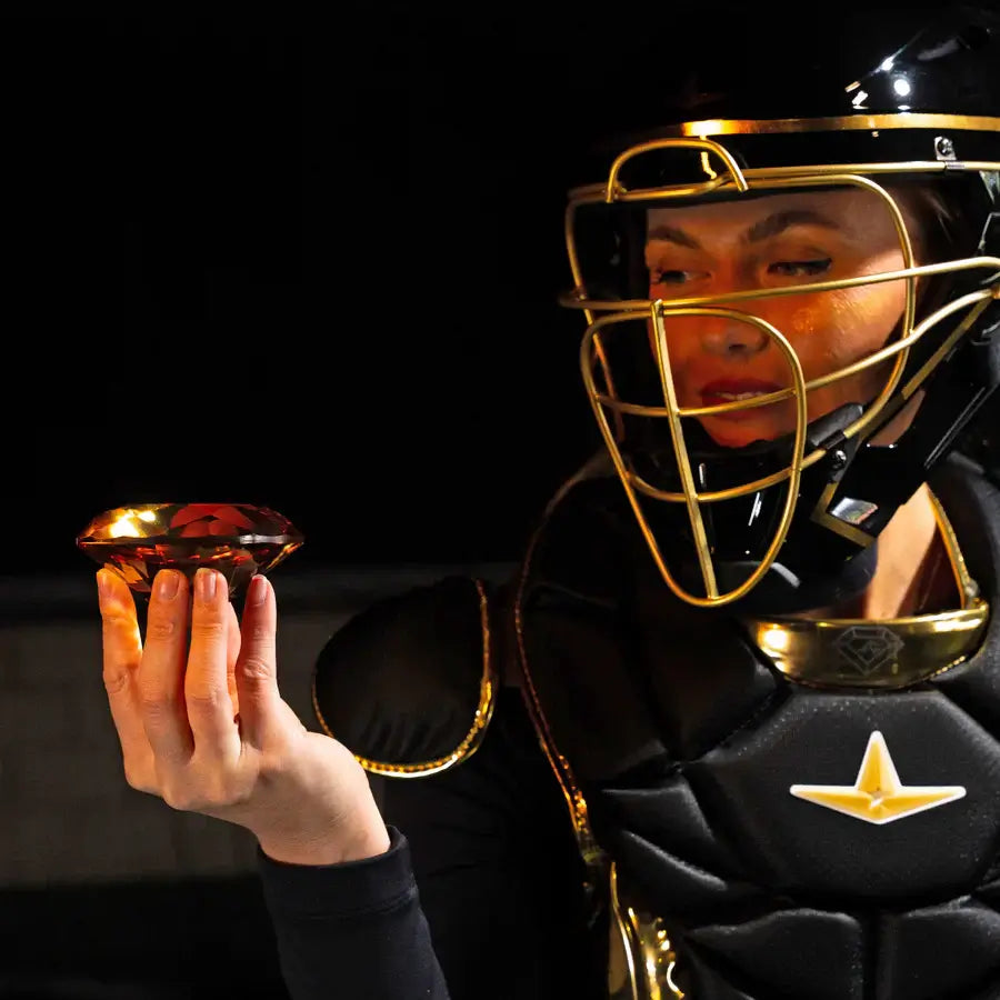 Catcher wearing All-Star PHX™ Diamond Series Softball Catcher's Kit Black/Gold, holding a gold diamond-shaped object.
