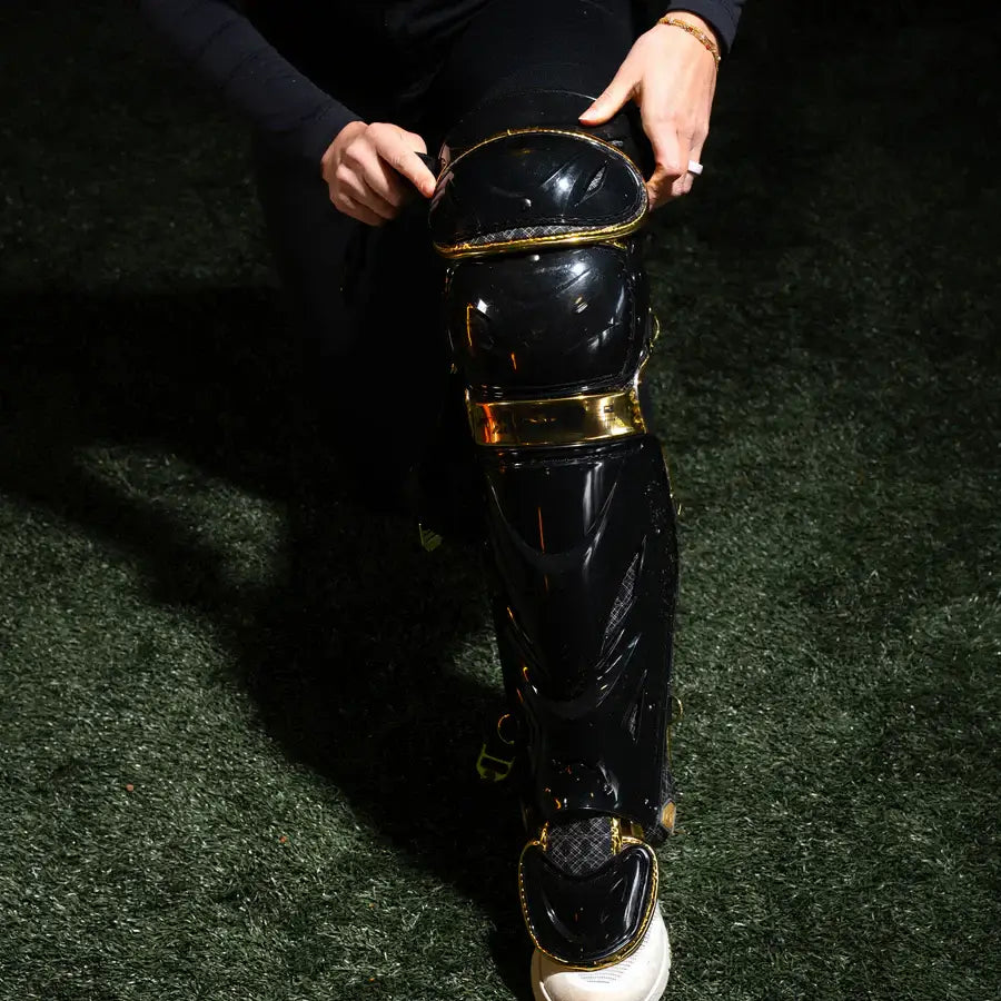 Applying All-Star PHX™ Diamond Series Softball Catcher's Kit leg guard in Black/Gold with detailed gold trim on knee and shin.