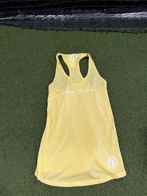 Yellow racerback women's t-shirt with "Between The Lines" printed on the front, laid on a green surface.